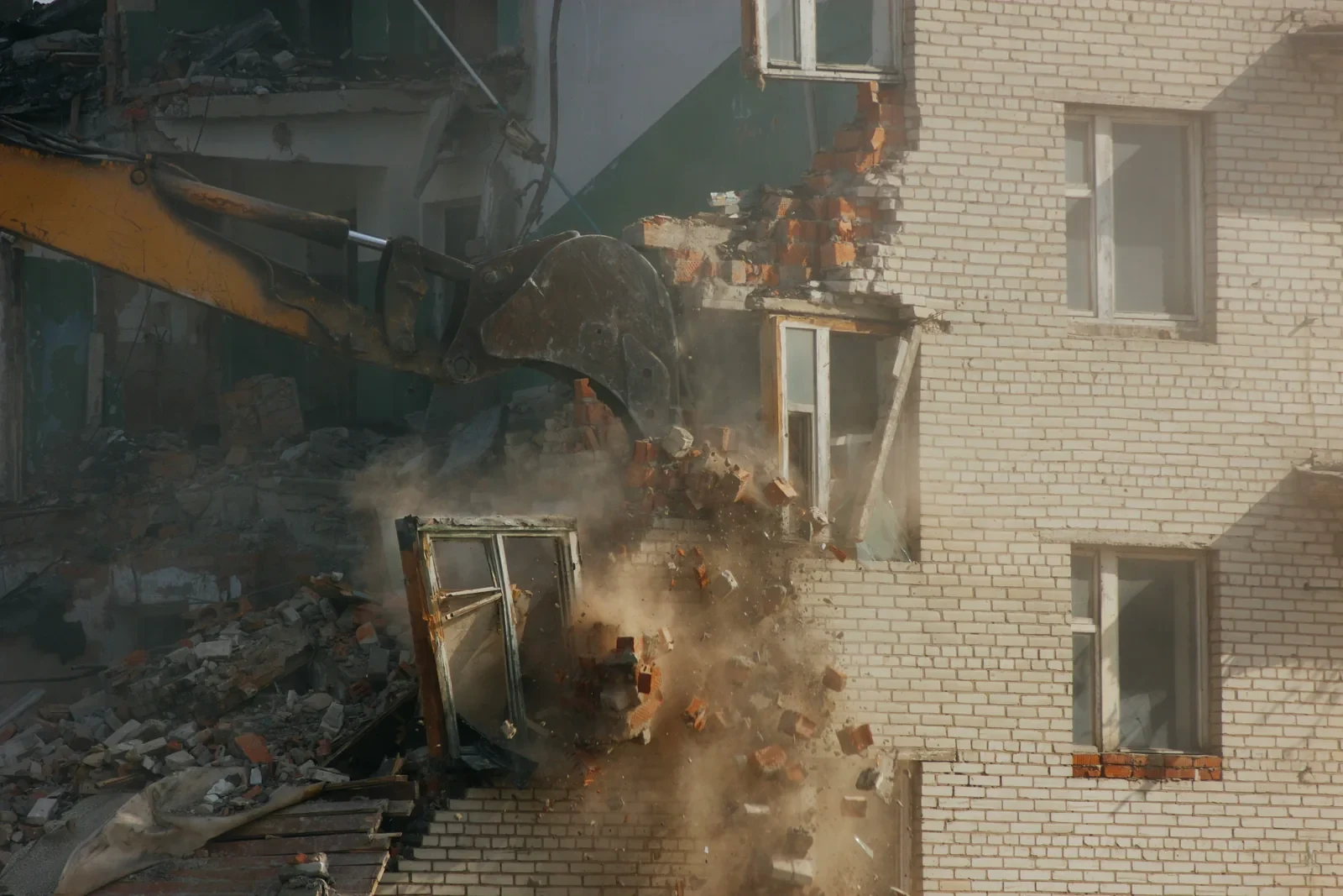 An excavator bucket knocking bricks out of a wall, dust and falling debris below