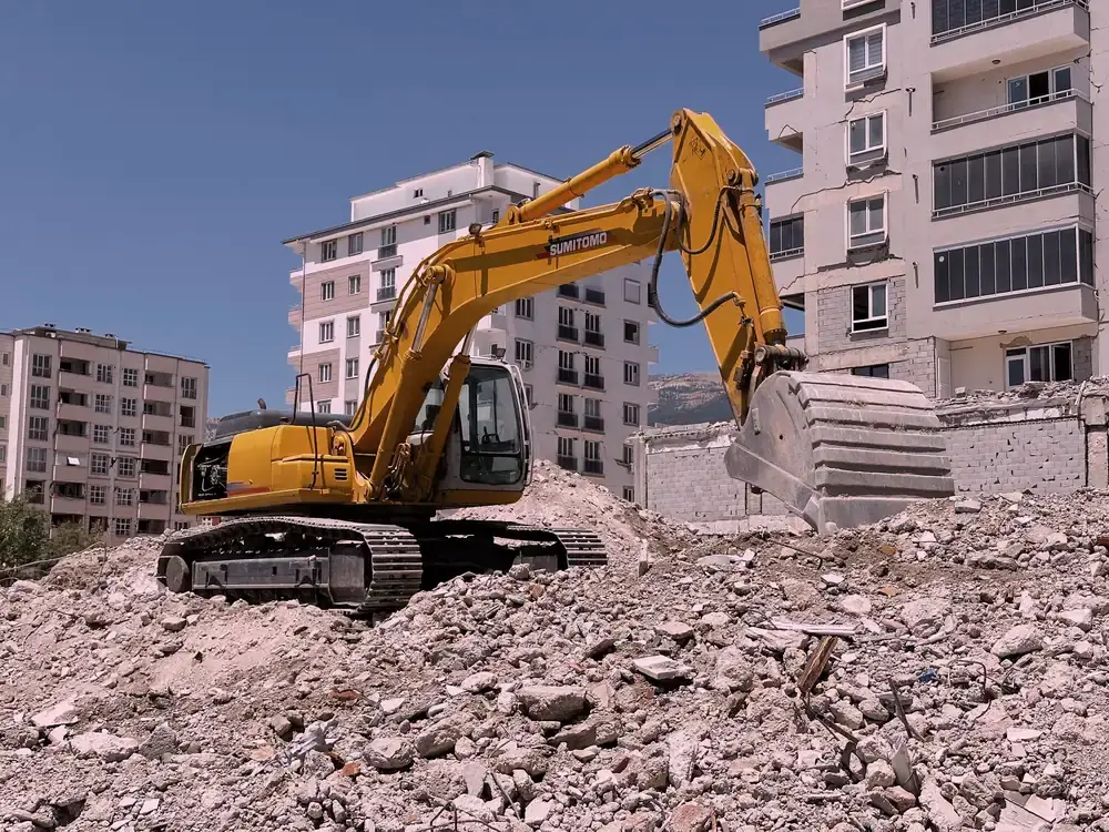 An excavator on a rubble pile in front of damaged concrete apartment blocks