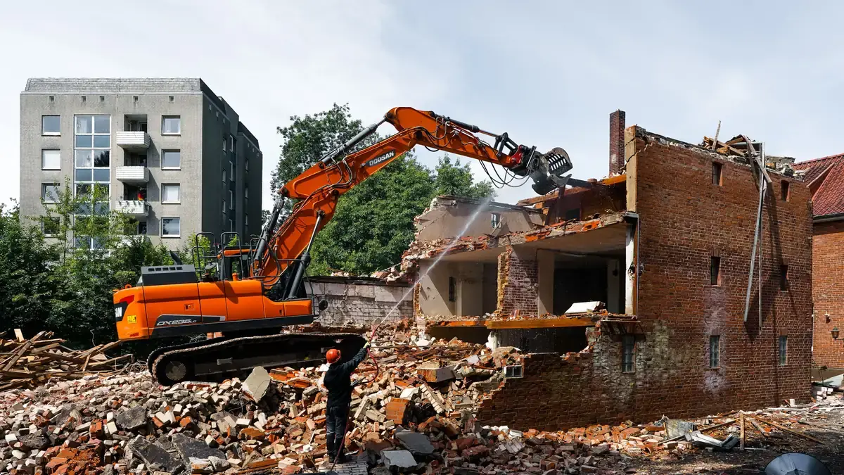 A worker damping down brick rubble with a hose while an excavator works behind